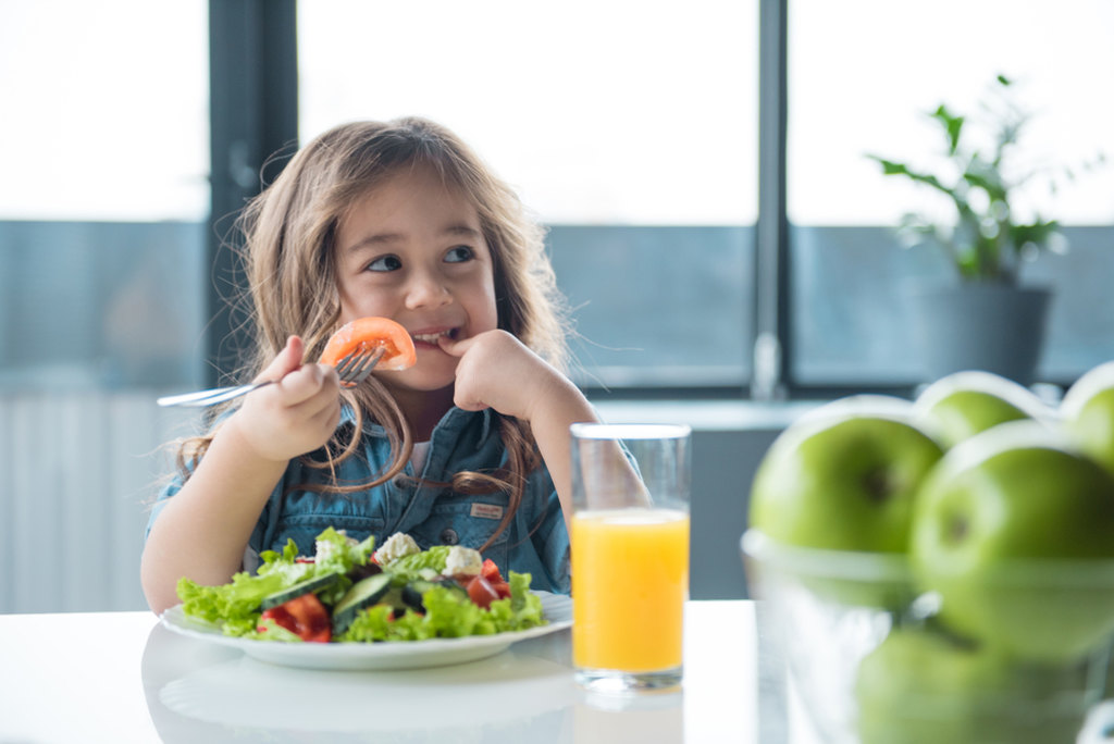 criança sentada na mesa comendo um prato com verduras e legumes e um copo de laranja para alimentação saudável infantil 